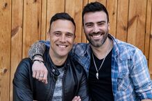 Dois homens sorridentes posam juntos em frente a um fundo de madeira