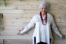 Mulher sorri com camisa branca e colar vermelho, junto a parede de madeira