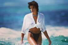 Mulher caminha na água com camisa branca e parte de baixo do biquíni preta