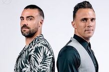 Dois homens posam juntos, um com camisa de padrão e outro com colete.
