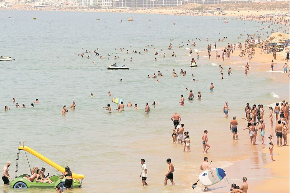 Praia lotada com banhistas e atividades aquáticas em dia soalheiro.
