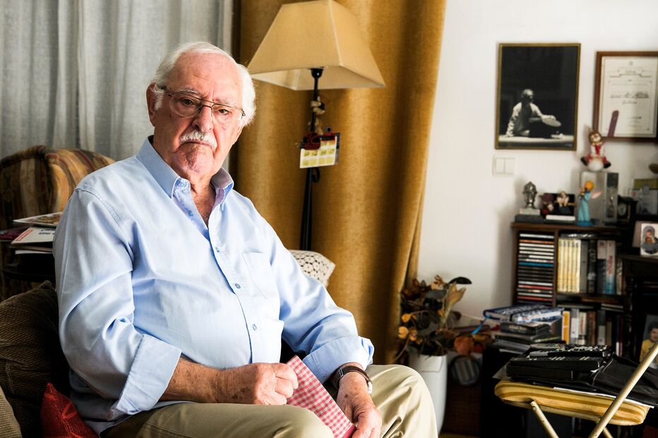 Homem mais velho sentado numa cadeira em casa com uma camisa azul e calças caqui