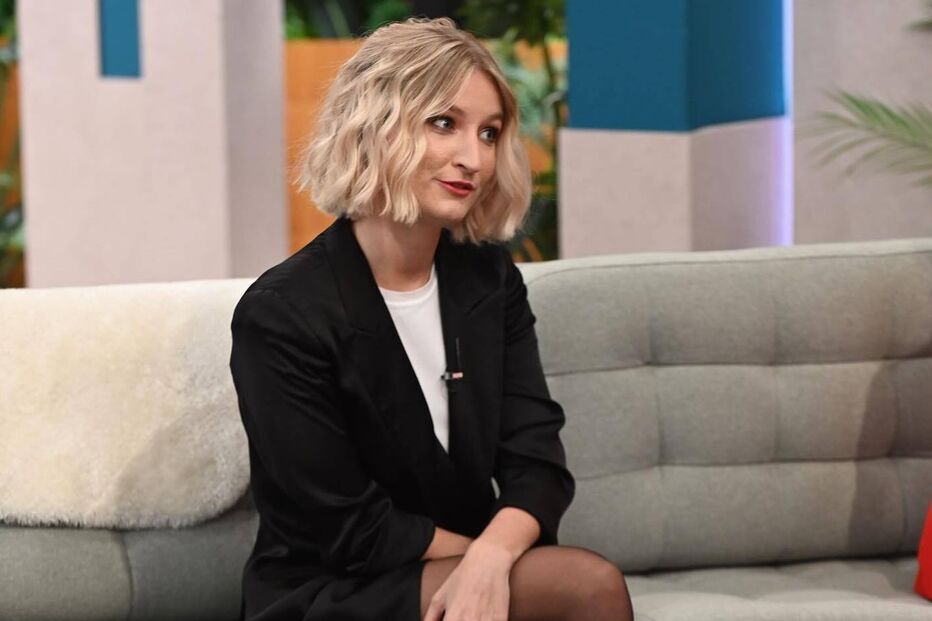 Mulher de cabelo louro encaracolado veste blazer preto e botas de cano alto num sofá