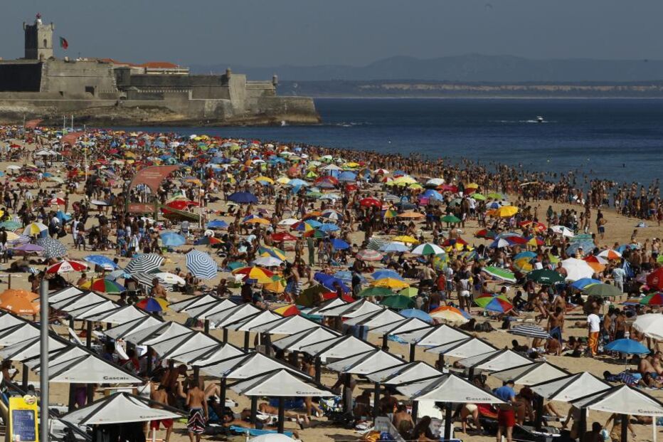 Praia da Carcavelos cheia de pessoas, com o Forte de São Julião da Barra ao fundo