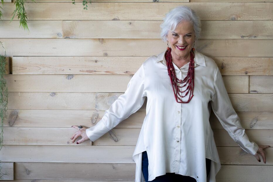 Mulher sorri com camisa branca e colar vermelho, junto a parede de madeira