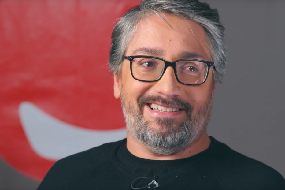 Ray Romano sorri com um fundo vermelho e usa óculos