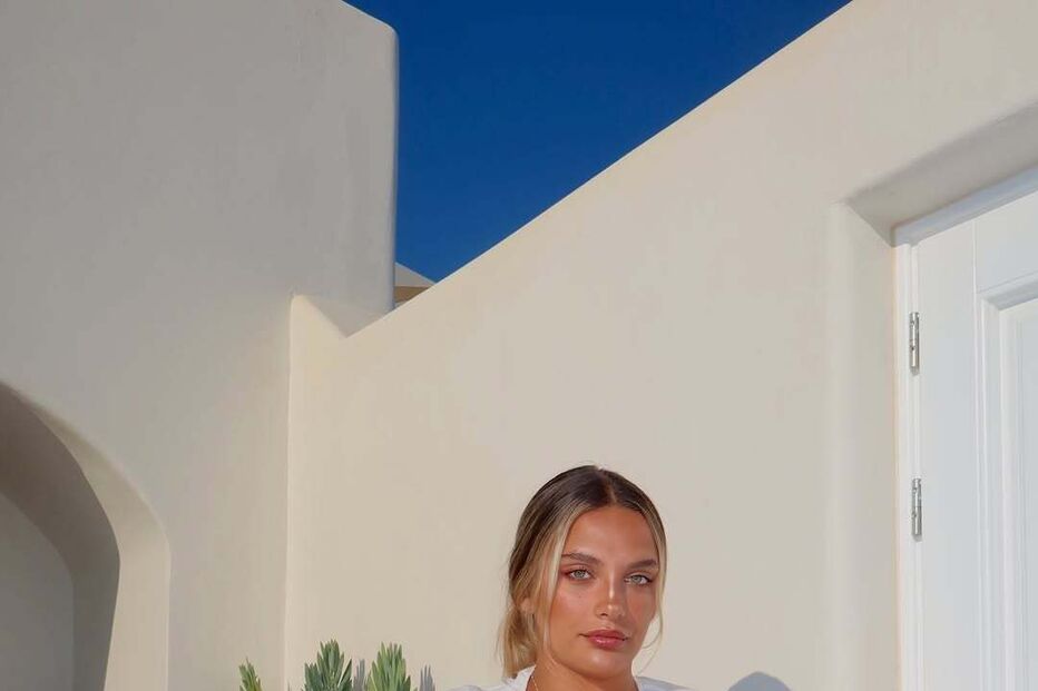 Mulher posa com conjunto branco e mala castanha num cenário de Santorini