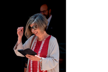 Mulher sorri e gesticula com um tablet na mão; ao fundo, um homem de barba observa