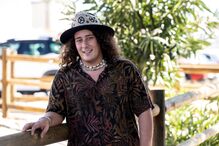 Homem sorri com camisa florida, chapéu estampado e colar de conchas