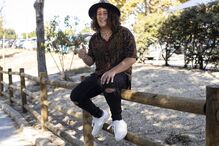 Jovem sorri, sentado numa cerca de madeira com um chapéu e roupa casual