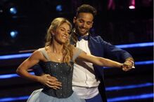 Casal dança num palco, a mulher usa um vestido azul de baile