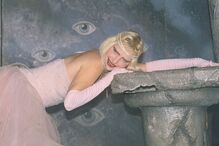 Mulher loira deitada com vestido rosa e luvas compridas, com maquilhagem brilhante