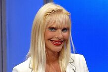 Mulher sorri com blazer branco, colar com cruz e um livro na mão