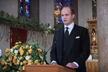 Homem discursa num funeral, com coroa de flores e igreja ao fundo