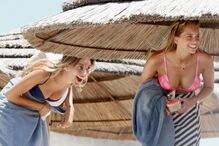 Duas mulheres sorriem na praia, sob um chapéu de sol