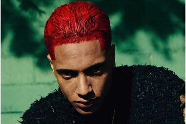 Homem com cabelo vermelho, colete preto e colar dourado posa para foto