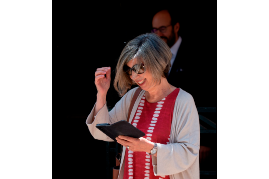 Mulher sorri e gesticula com um tablet na mão; ao fundo, um homem de barba observa