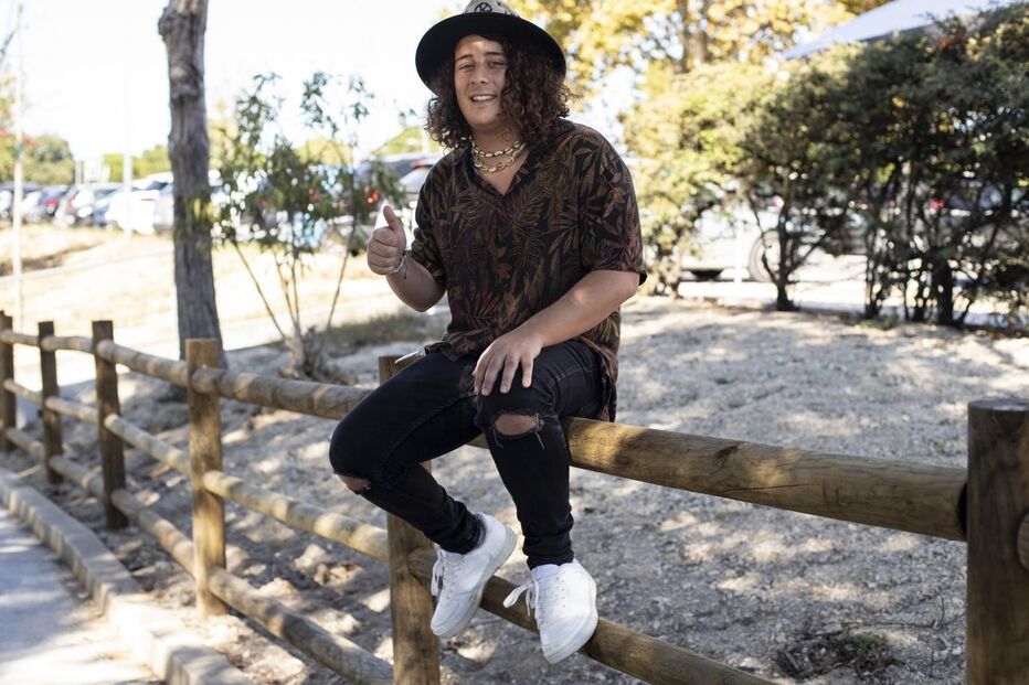 Jovem sorri, sentado numa cerca de madeira com um chapéu e roupa casual