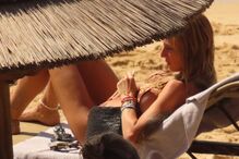 Mulher relaxa na praia sob um chapéu de sol, com acessórios e tatuagens visíveis