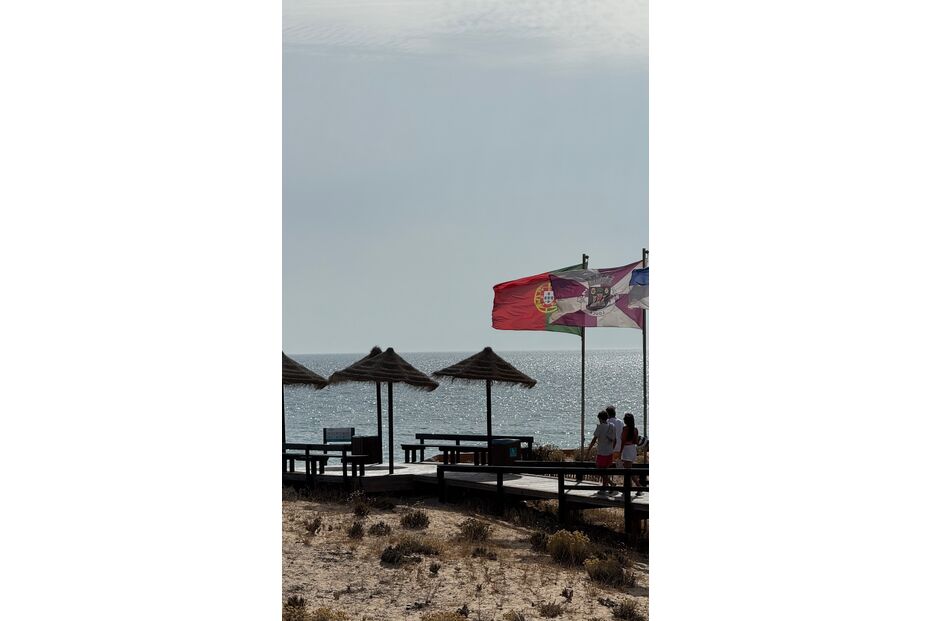 Praia com bandeiras hasteadas, passadiço de madeira e pessoas a passear