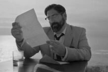 Homem de óculos lê carta à beira-mar num cenário a preto e branco