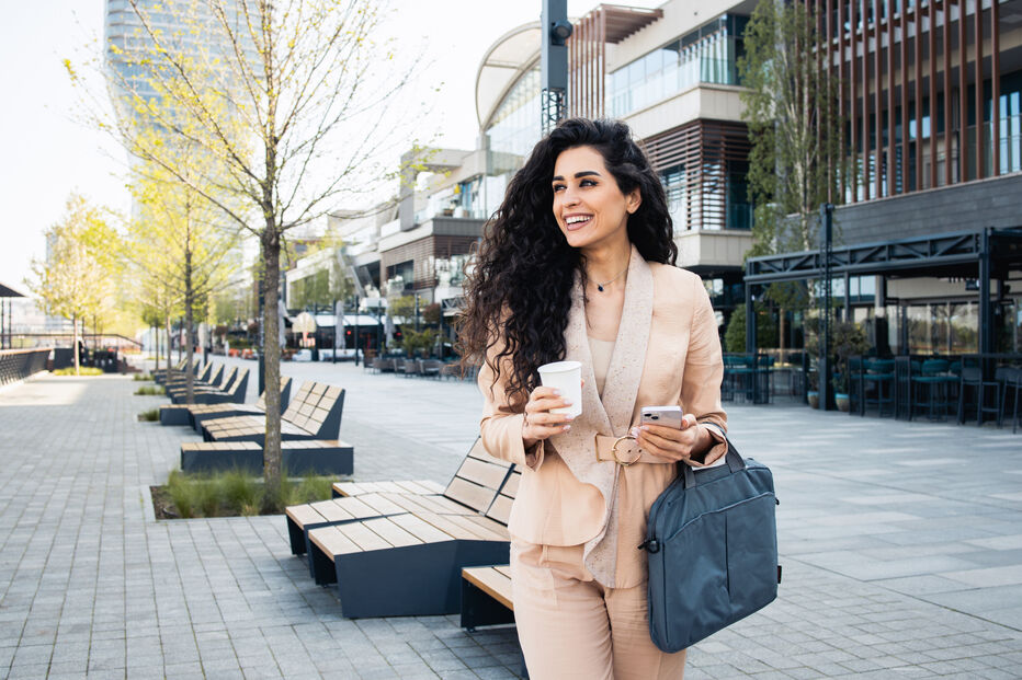 Mulher sorri na rua, com café e telemóvel, a caminho do trabalho