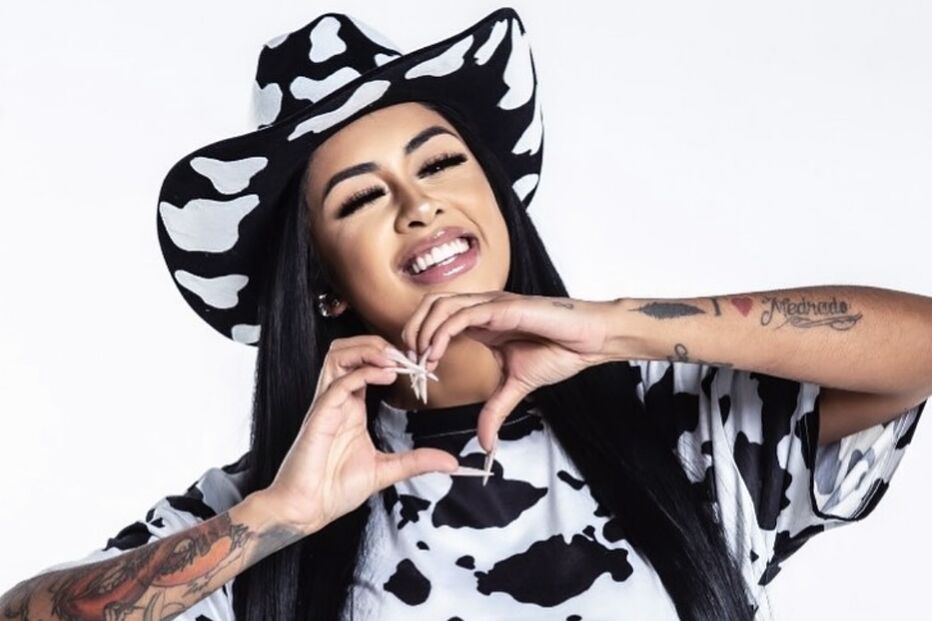 Mulher sorri com chapéu e roupa com padrão de vaca, e faz um coração com as mãos