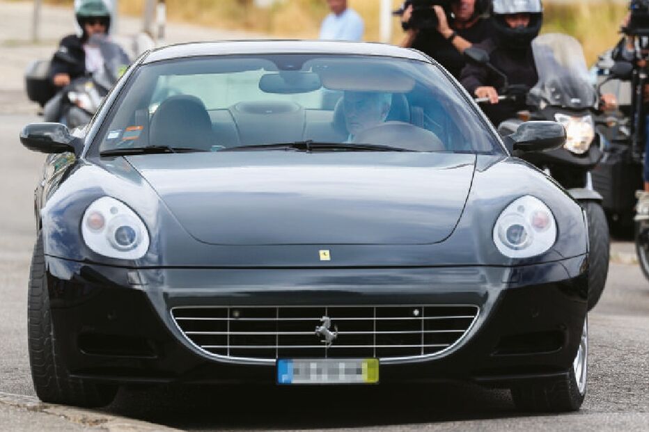 Um carro Ferrari preto seguido por fotógrafos e curiosos