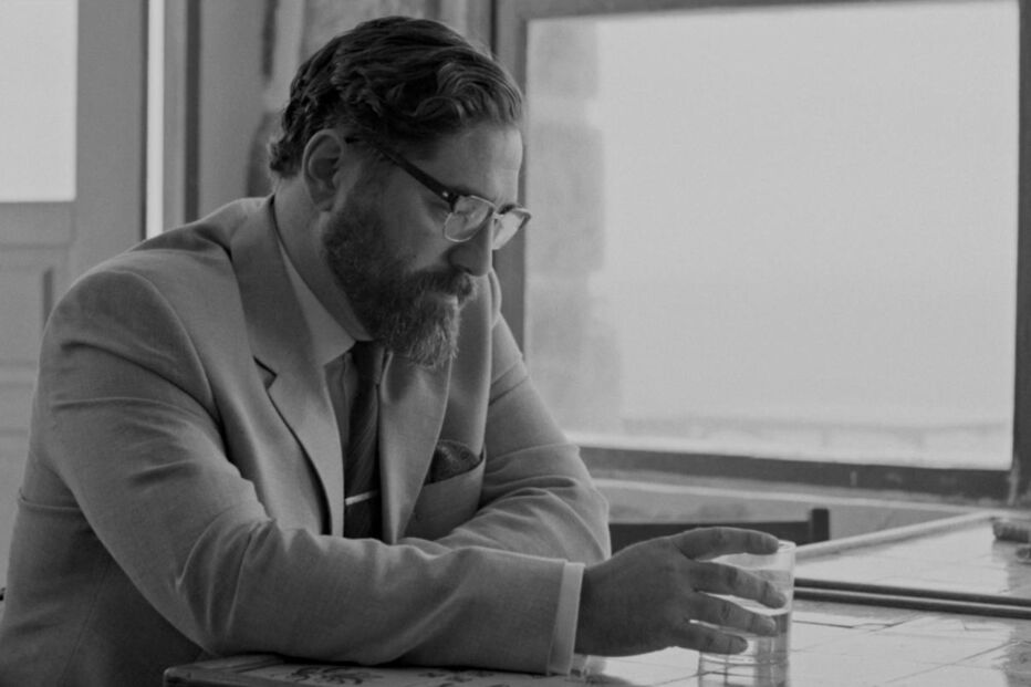 Um homem de fato e gravata, óculos e barba pousa a mão num copo