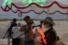 Músicos atuam na praia com violino e guitarra sob luzes coloridas