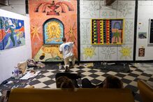 Artista pinta mural numa sala, com visitantes sentados