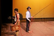 Dois homens em palco interagem com adereços