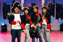 Grupo posa com trajes militares e guitarra num palco iluminado