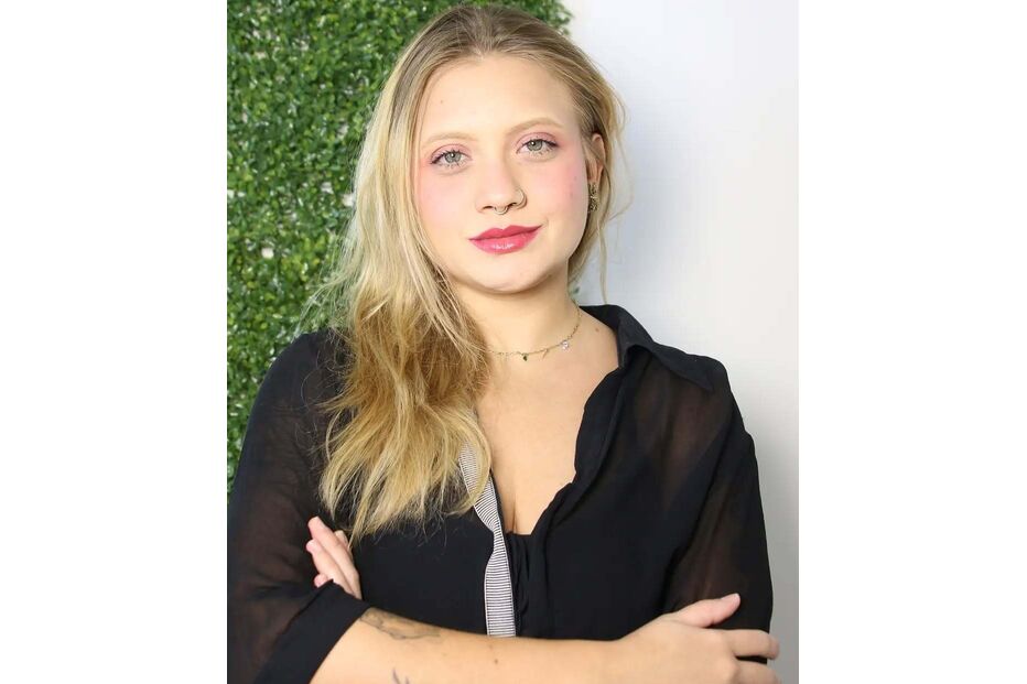 Mulher loira posa com camisa preta, braços cruzados e tatuagem