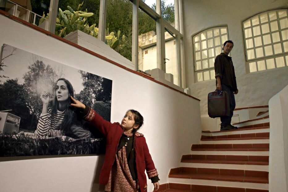 Uma menina aponta para fotografia enquanto um homem sobe as escadas com mala
