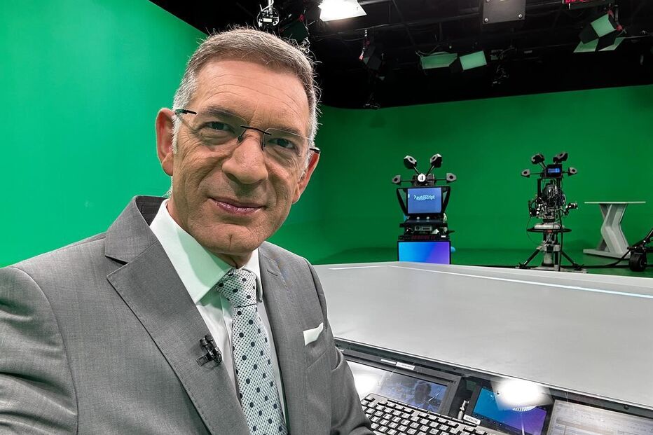 Um homem sorri num estúdio de televisão com fundo verde e equipamento profissional.
