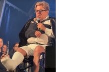 Homem discursa em palco de cadeira de rodas, com braço e perna engessados