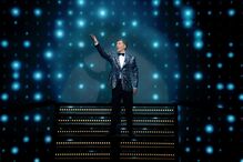 João Baião sobe ao palco, sorrindo e acenando para o público.
