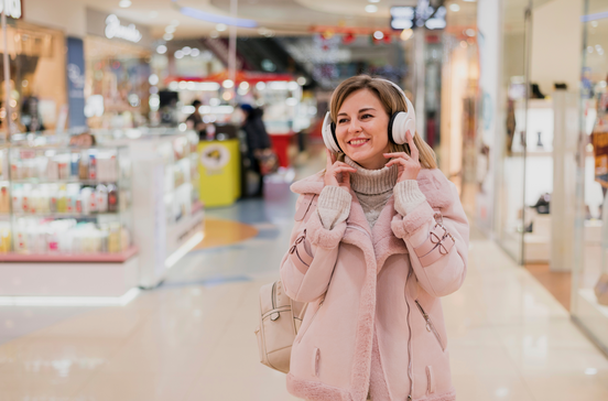 Mulher sorri num centro comercial com auscultadores e casaco rosa, aproveitando ofertas em tecnologia