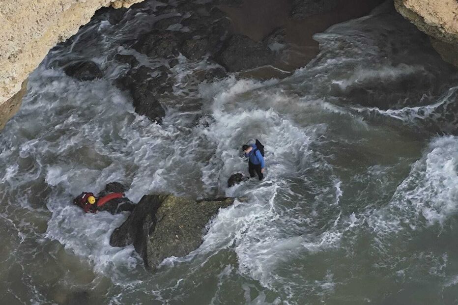 Duas pessoas na água, uma flutuando e outra caminhando perto de rochas na costa