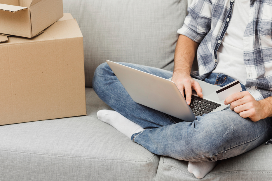Homem faz compras online com cartão e computador, sentado num sofá, junto a caixas