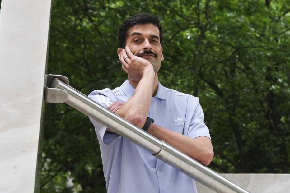 Homem posa encostado a estrutura metálica, com árvores verdes no fundo