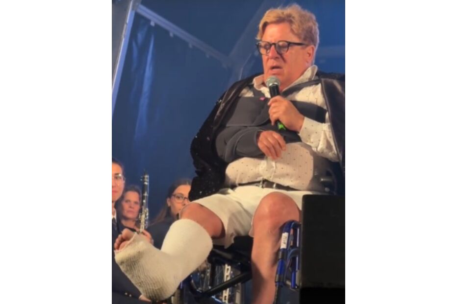 Homem discursa em palco de cadeira de rodas, com braço e perna engessados