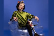 Mulher sentada numa cadeira transparente com vestido verde, prótese na perna e bota com padrão leopardo