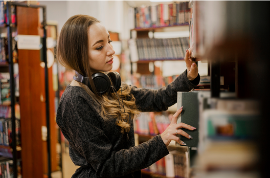 Mulher pesquisa livros numa biblioteca com auscultadores ao pescoço