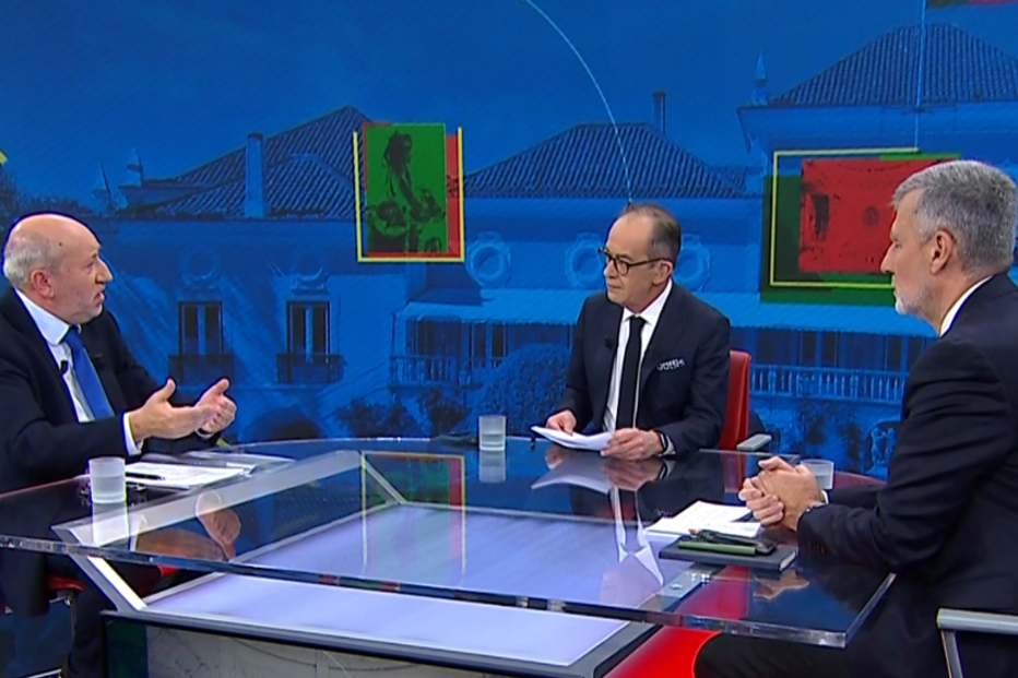 Três homens em debate televisivo num estúdio com o Palácio da Ajuda como fundo