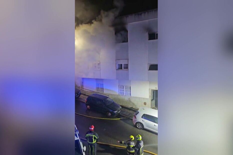 Bombeiros combatem incêndio num edifício; fumo visível e carros estacionados na rua