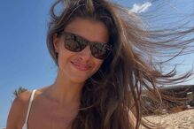 Mulher sorri na praia com óculos de sol, cabelo ao vento e céu azul