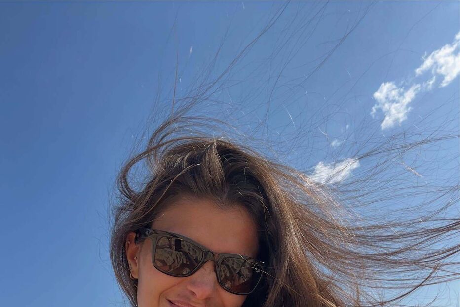 Mulher sorri na praia com óculos de sol, cabelo ao vento e céu azul