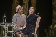 Espetáculo teatral com dois atores, mesa e champanhe em palco.
